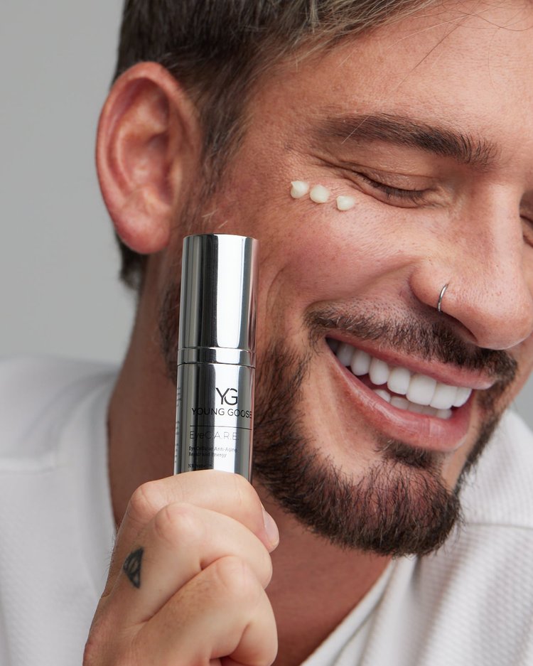 Smiling man holding Young Goose EyeC.A.R.E. NAD+ Boosting Eye Cream with a dab of cream on his cheek